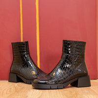 Black crocodile-patterned boots on a wooden floor with a red and yellow striped wall in the background.